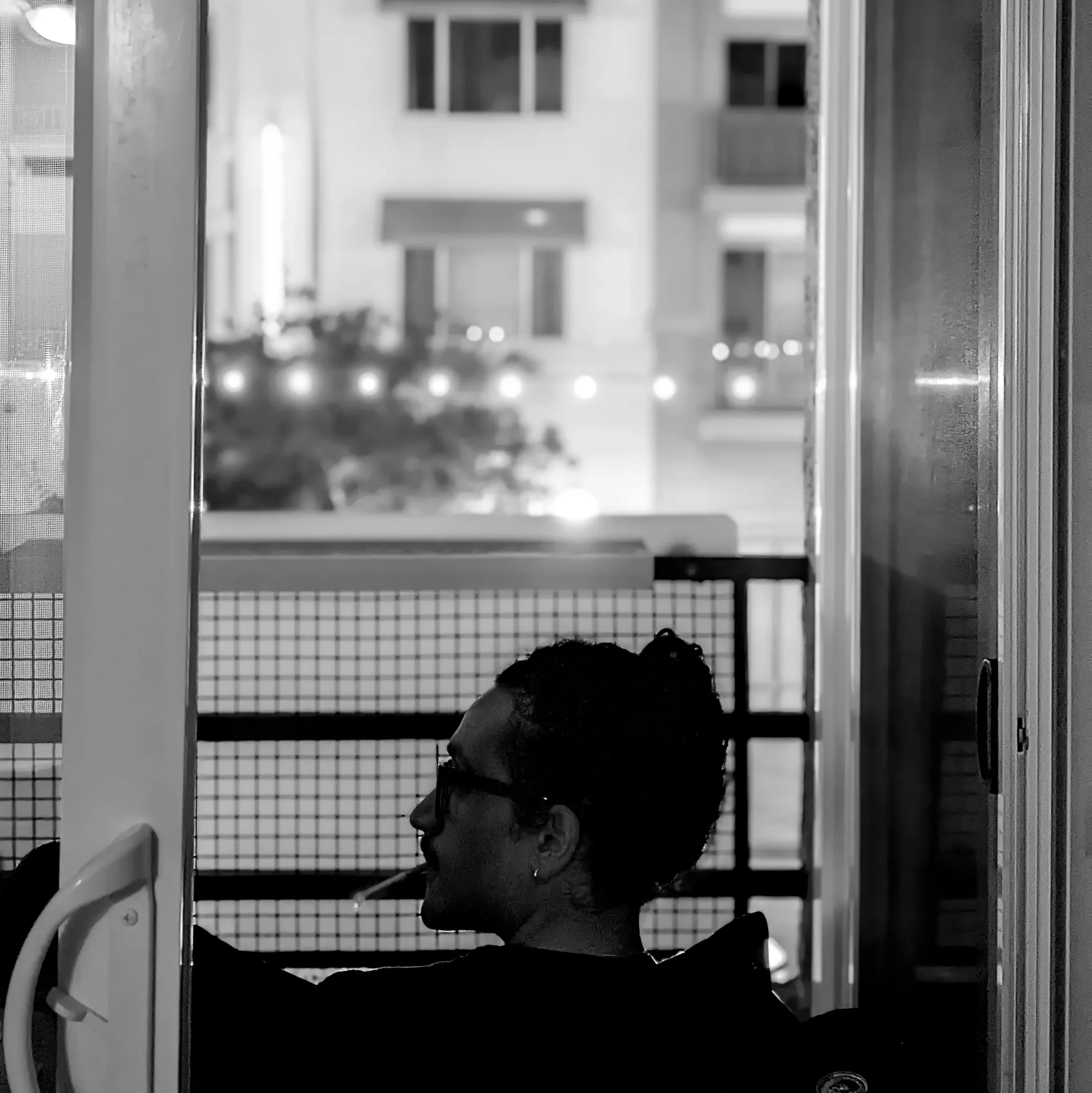 A black-and-white portrait of someone in glasses smoking on a balcony, framed by an open door