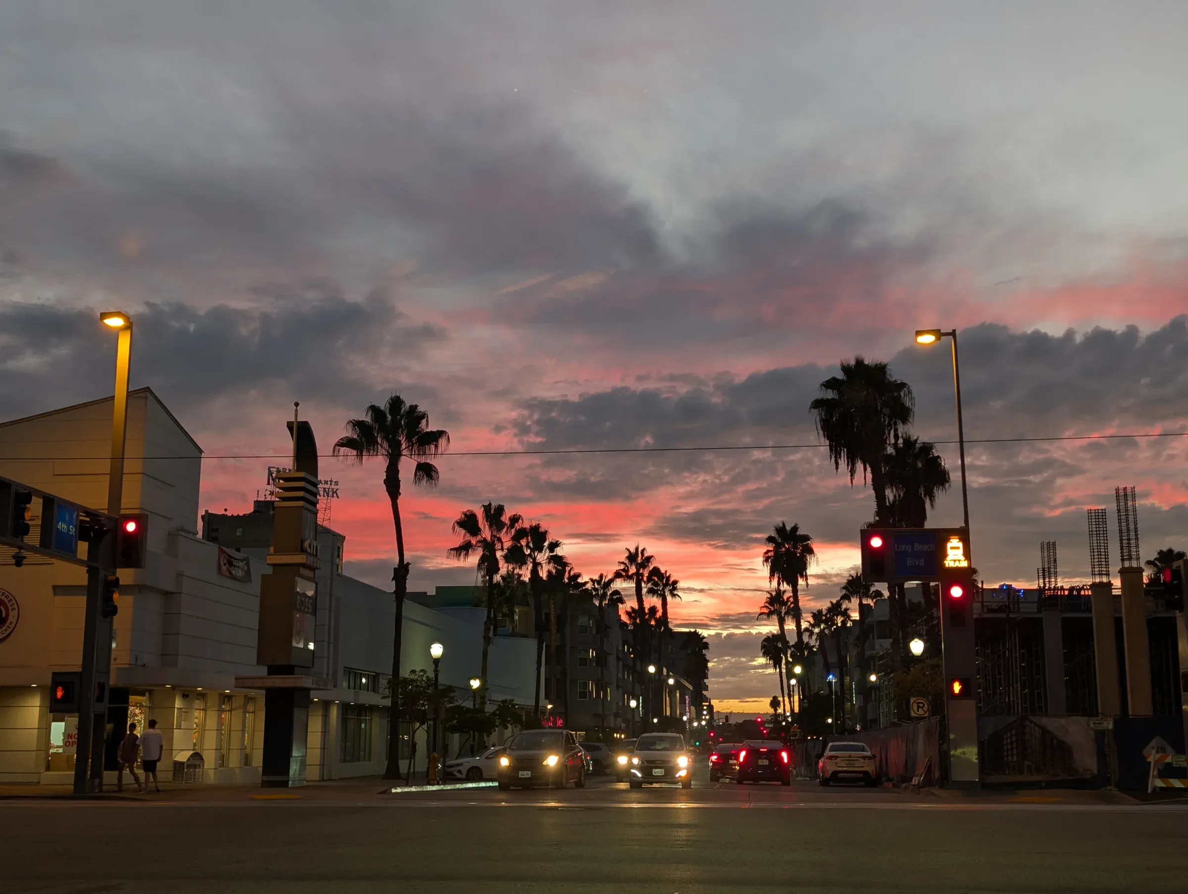 A wide street at dusk lined with palm trees and streetlights, under a pink and gray sky