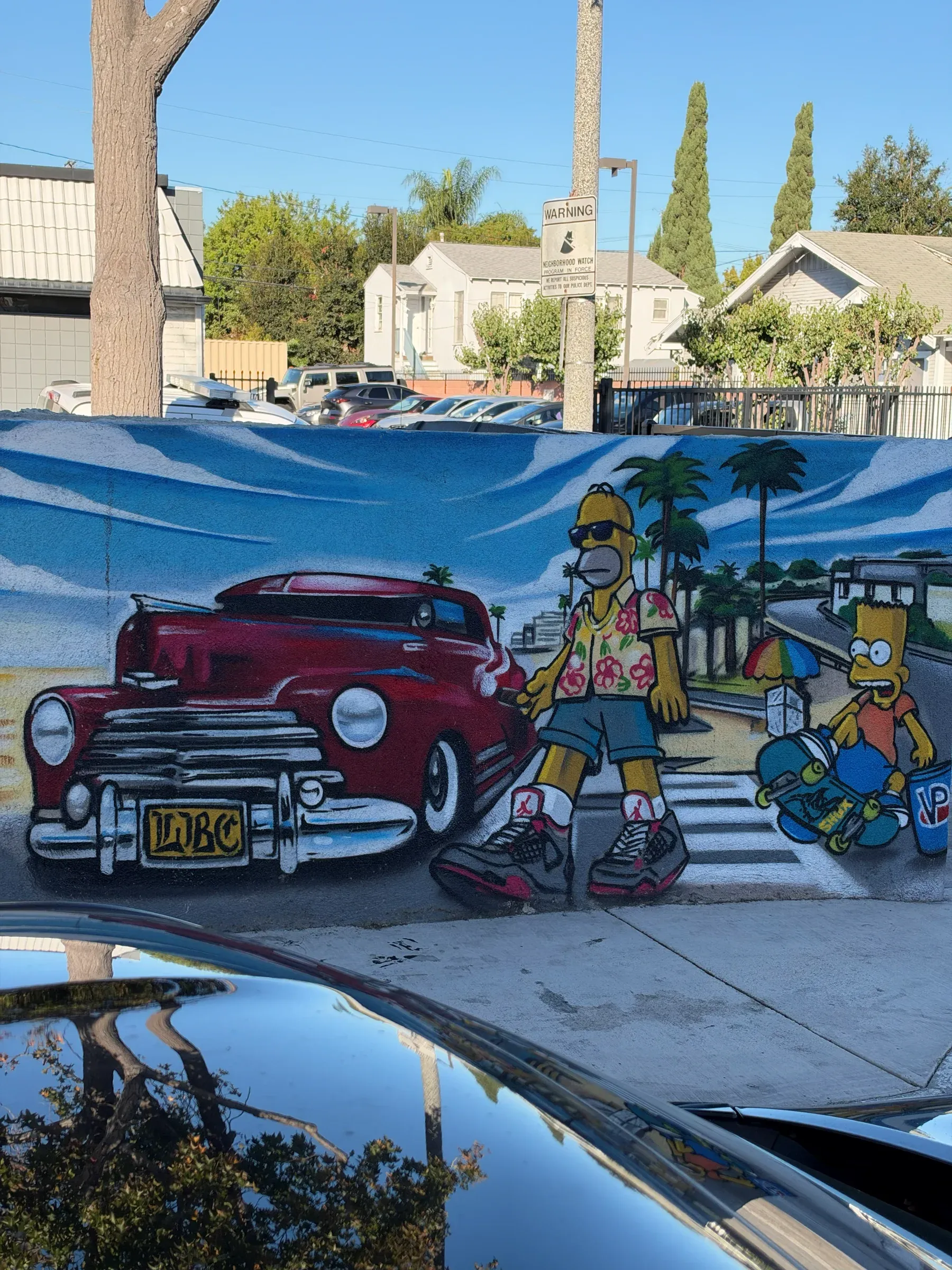 A street mural of Homer and Bart Simpson walking past a red lowrider with an LBC plate, under a blue sky
