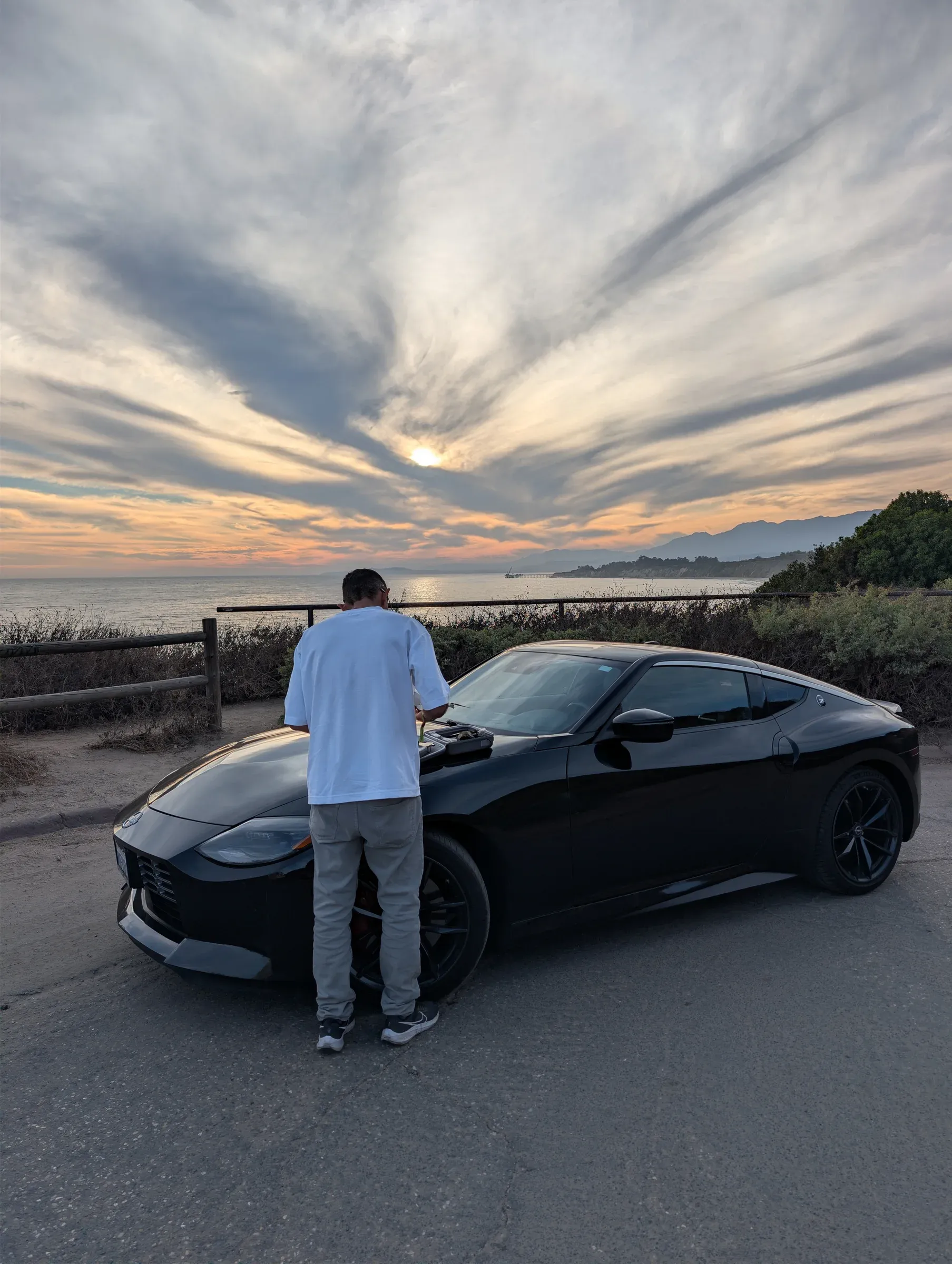 Someone in a white T-shirt leaning over a black sports car at an overlook by the sea, under streaked clouds at sunset