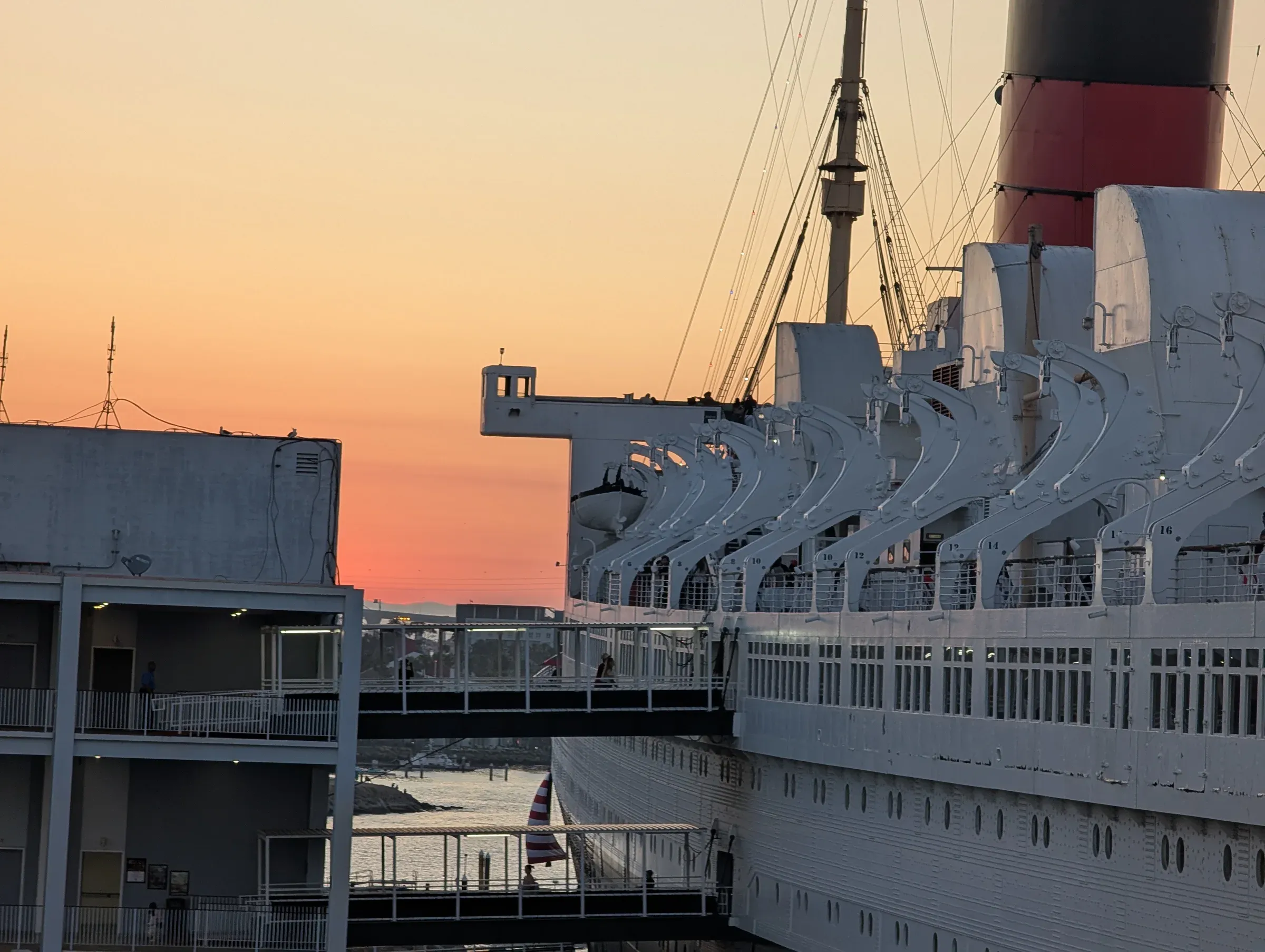 The side of the Queen Mary, its lifeboat davits and red funnel against an orange sunset