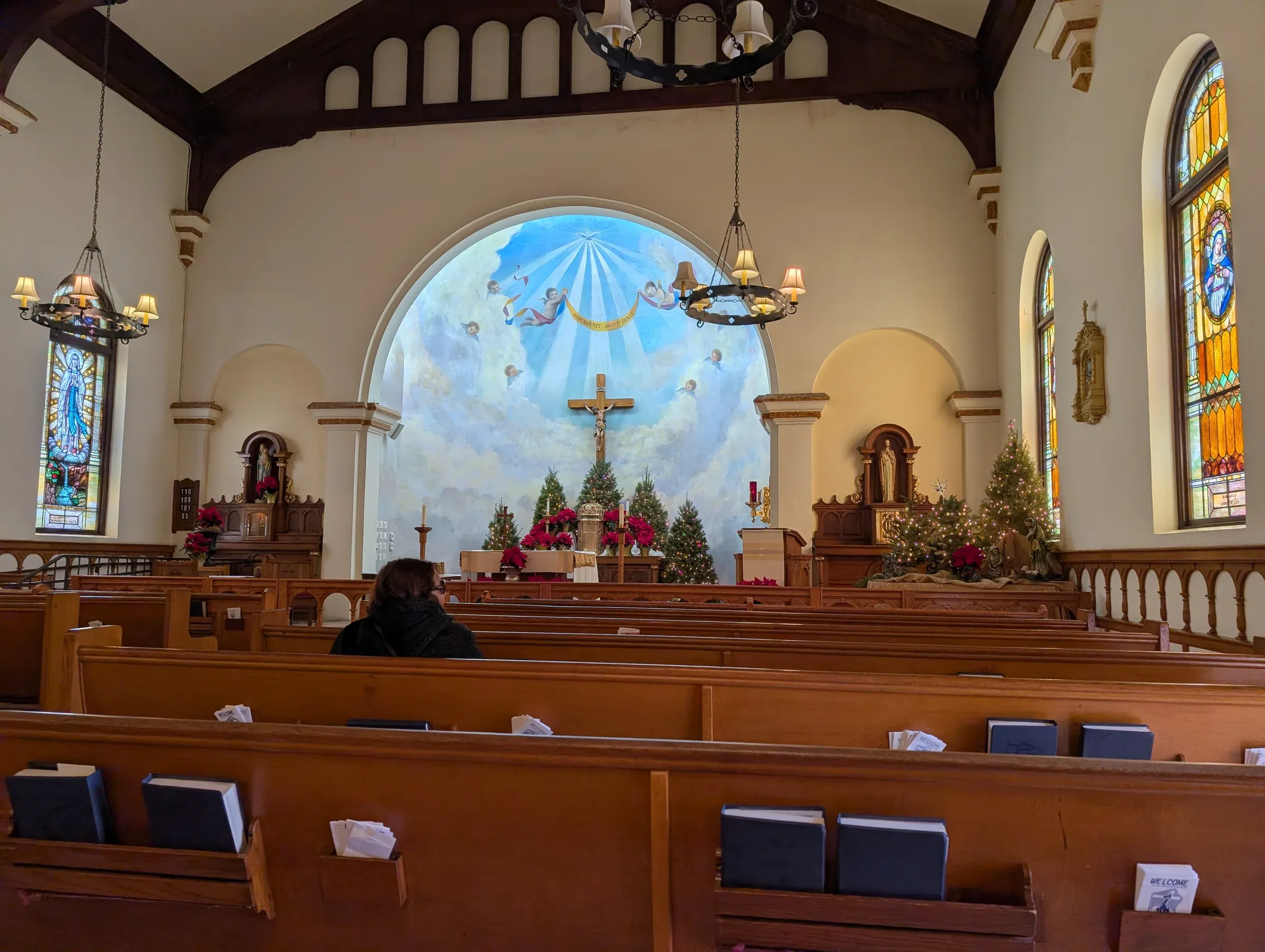 Inside a quiet church with wooden pews, stained glass and a painted sky behind the altar, where one person sits alone