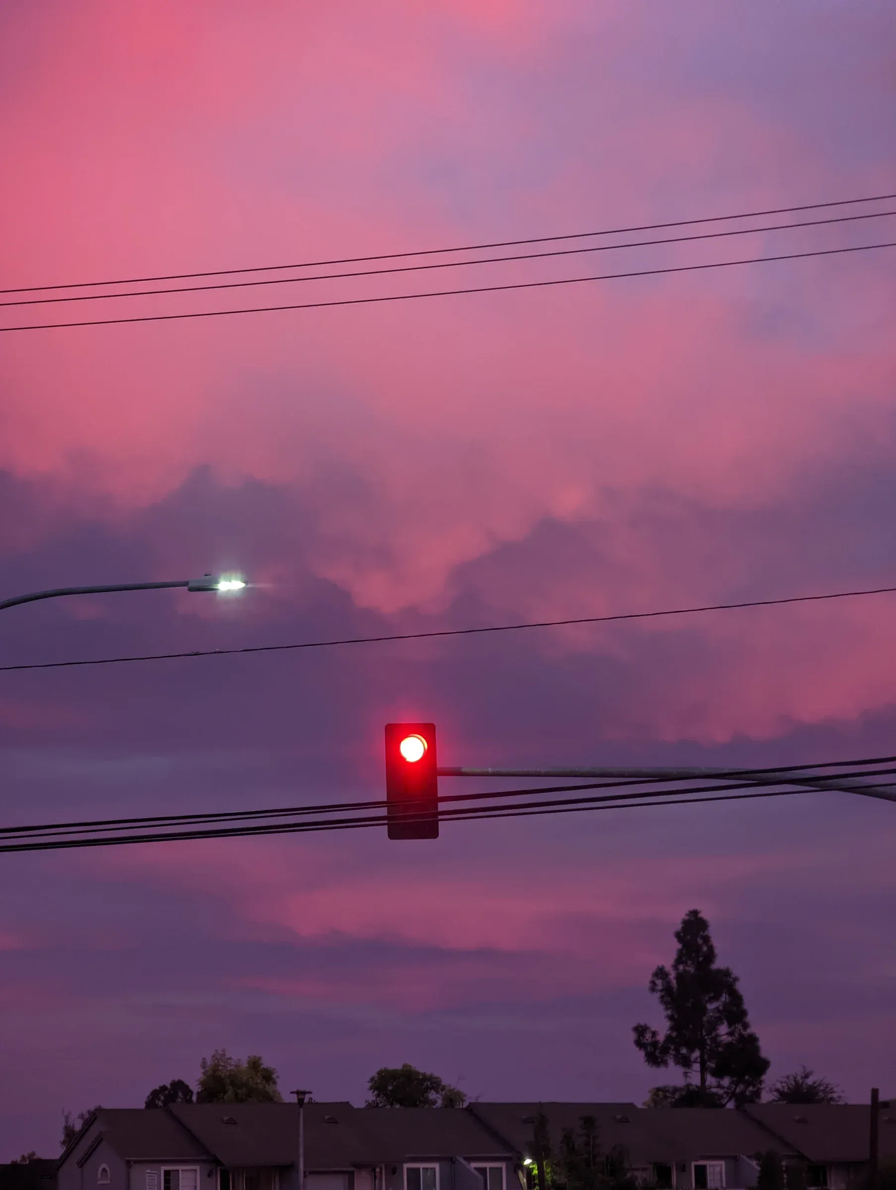 A red traffic light and power lines against a pink and purple evening sky over rooftops